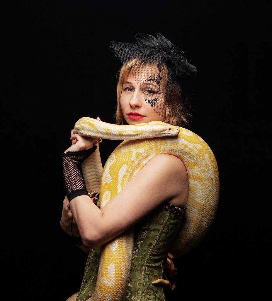 Side view of young lady in stylish hat with veil and mesh gloves holding yellow anaconda on shoulders while standing on black background and looking at camera